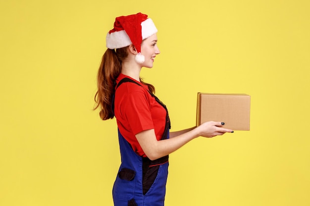 Vue latérale portrait d'une femme de messagerie remettant un colis en carton pour le client, présente pour le Nouvel An, portant une salopette bleue et un chapeau de père noël. Studio intérieur tourné isolé sur fond jaune.