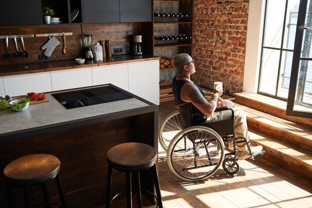 Vue latérale portrait d'une femme tatouée contemporaine en fauteuil roulant savourant un café à la maison et regardant la fenêtre, espace pour copie