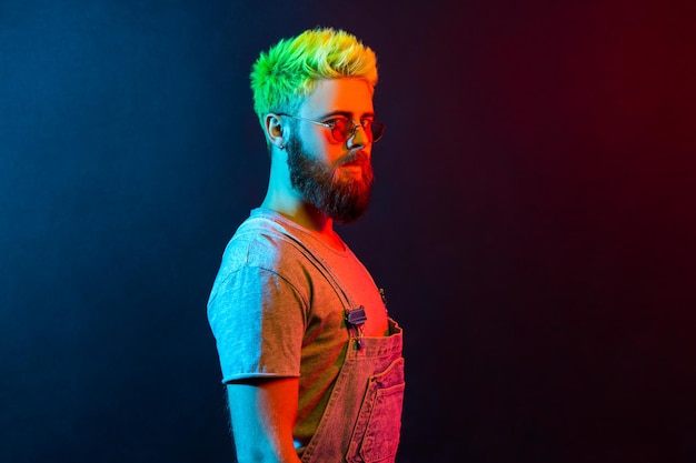 Vue latérale portrait d'un gars avec une barbe et des cheveux verts regardant la caméra avec une expression sérieuse et confiante portant une salopette en jean copie espace Néon coloré tourné en studio à l'intérieur