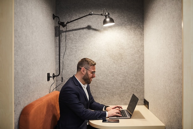 Vue latérale portrait d'homme d'affaires barbu souriant à l'aide d'un ordinateur portable tout en appréciant le travail dans la cabine de café au bureau, copiez l'espace