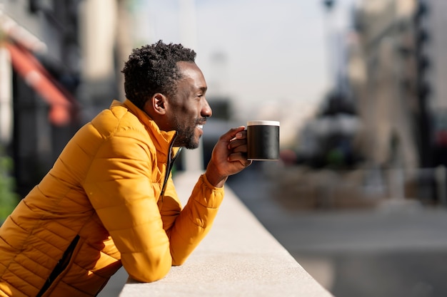 Vue latérale portrait d'un homme africain souriant appuyé sur un balcon tenant une tasse de café en contemplant des vues dans une ville