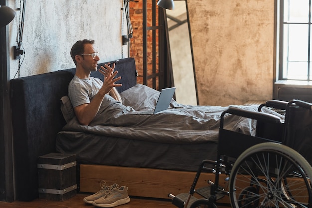 Vue latérale portrait d'un homme handicapé moderne parlant par smartphone au lit, fauteuil roulant au premier plan, espace pour copie