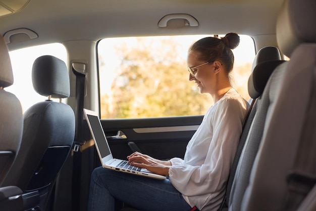 Vue latérale portrait d'une jeune femme d'affaires utilisant un ordinateur portable assis sur le siège arrière de la voiture en tapant sur un ordinateur portable en tapant un document important fonctionne en ligne pendant un voyage