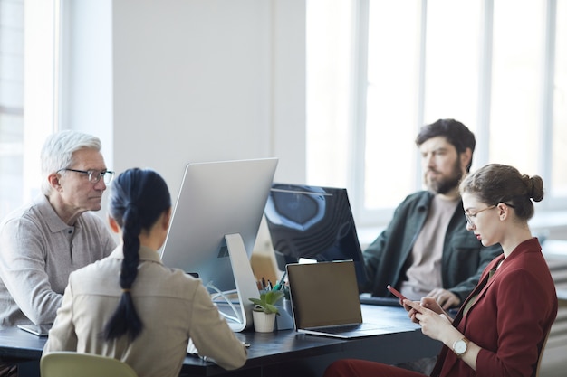 Vue latérale portrait d'une jeune femme élégante utilisant un smartphone tout en travaillant avec divers groupes de personnes dans un bureau moderne