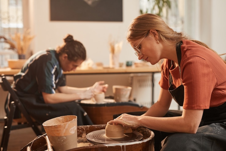 Photo vue latérale portrait d'une jeune femme façonnant de l'argile sur un tour de poterie tout en faisant de la céramique dans un atelier éclairé par la lumière du soleil, espace pour copie
