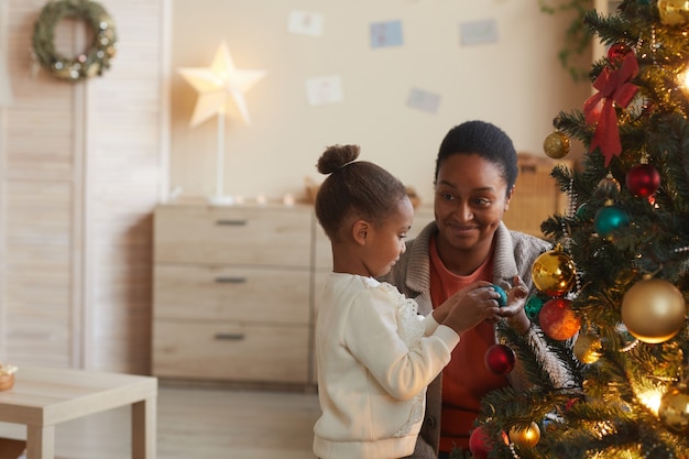 Vue latérale portrait de jolie fille afro-américaine décoration arbre de Noël avec maman heureuse souriante dans un intérieur confortable, espace copie