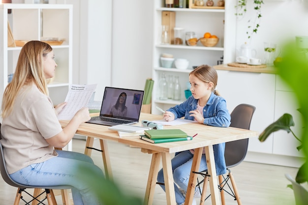 Vue latérale portrait de mère à faire ses devoirs avec sa jolie fille assise au bureau et regarder des vidéos de l'école en ligne, espace copie