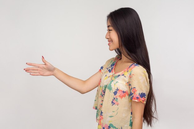 Vue latérale d'une sympathique fille asiatique aux longs cheveux bruns en chemisier donnant la main à la poignée de main et souriant au client saluant et faisant connaissance en studio intérieur isolé sur fond gris