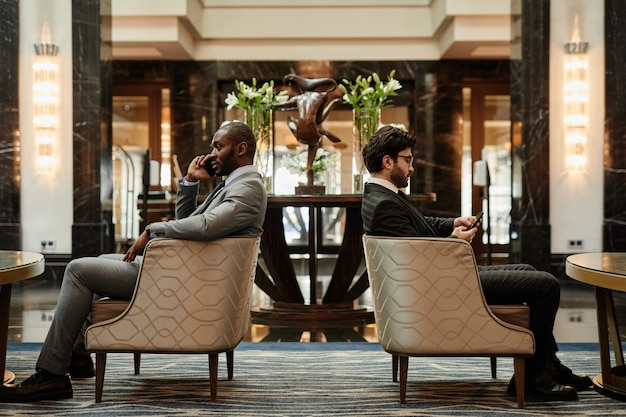 Vue latérale sur toute la longueur de deux hommes d'affaires prospères se relaxant dans des fauteuils dans le hall de l'hôtel luxueux