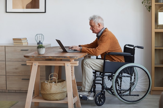 Vue latérale sur toute la longueur d'un homme âgé en fauteuil roulant utilisant un ordinateur portable à la maison, espace pour copie