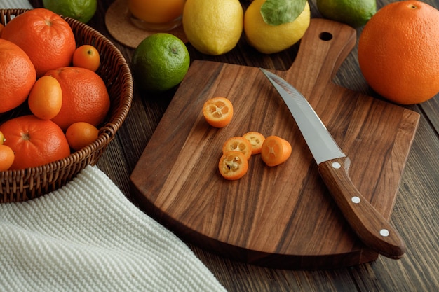 Vue latérale des tranches de kumquat avec un couteau sur une planche à découper et des mandarines dans le panier citron citron vert orange sur fond de bois