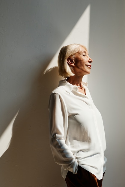 Vue latérale verticale portrait d'une femme mûre élégante éclairée par la lumière du soleil contre l'espace de copie de mur blanc