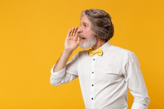 Vue latérale d'un vieil homme barbu moustache aux cheveux gris en chemise blanche noeud papillon posant isolé sur fond jaune. Concept de style de vie des gens. Maquette de l'espace de copie. Crier avec un geste de la main près de la bouche.