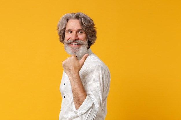 Vue latérale d'un vieil homme barbu à moustache aux cheveux gris souriant en chemise blanche noeud papillon posant isolé sur fond jaune en studio. Concept de style de vie des gens. Maquette de l'espace de copie. Pouce pointé de côté.