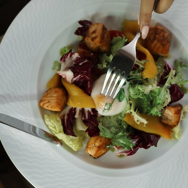 Vue Sur Les Mains Avec Des Couverts Et Salade De Légumes Et œuf Poché Sur Une Plaque Blanche à L'intérieur Sur La Table. Fourchette Et Couteau Entre Les Mains De La Jeune Fille. Petit-déjeuner Sain