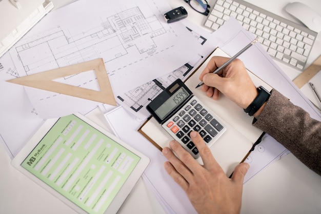 Vue des mains de l'ingénieur avec stylo et calculatrice prendre des notes dans le cahier tout en travaillant au bureau au bureau