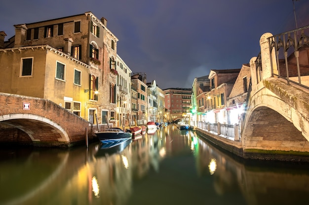 Vue nocturne de vieux bâtiments illuminés, de bateaux flottants et de reflets lumineux dans l'eau du canal à Venise, en Italie.