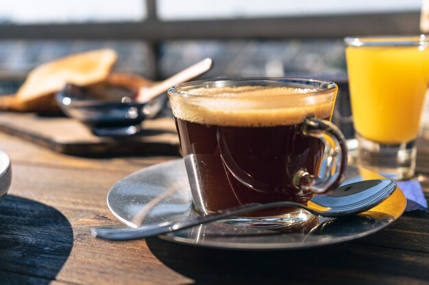 Vue normale d'une tasse de café noir au premier plan, avec un jus d'orange et des toasts derrière.