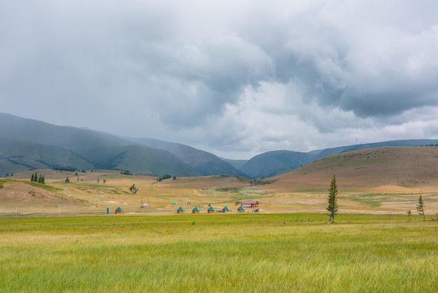 Vue panoramique depuis la steppe herbeuse ensoleillée jusqu'aux petites maisons sur fond de haute chaîne de montagnes verte dans des nuages pluvieux Paysage spectaculaire avec prairie et petits lodges dans les montagnes par temps variable