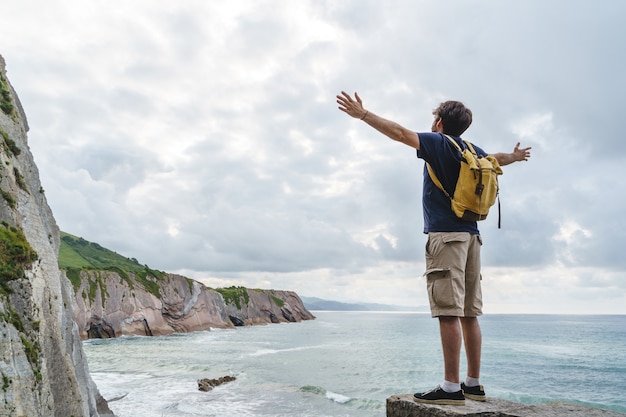 Vue panoramique d'un homme méconnaissable aux bras grands ouverts dans une falaise. Vue horizontale du routard voyageant à l'extérieur avec l'océan bleu en arrière-plan.