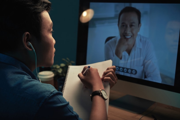 Vue par-dessus l'épaule d'un étudiant asiatique dans des écouteurs regardant un webinaire et prenant des notes dans un carnet de croquis à n...