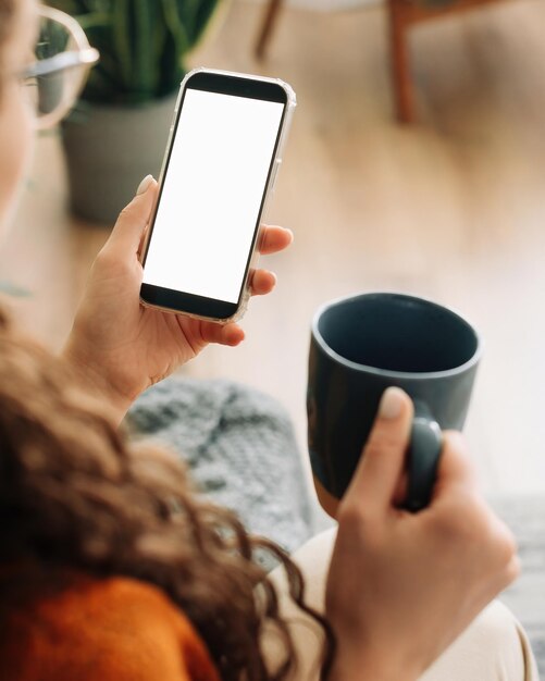 Vue par-dessus l'épaule d'une femme tenant un café et un smartphone avec un écran cellulaire de maquette blanche dans les mains