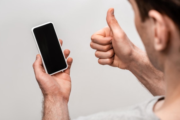 Vue partielle de l'homme montrant le pouce vers le haut tout en tenant un smartphone avec écran blanc isolé sur gris