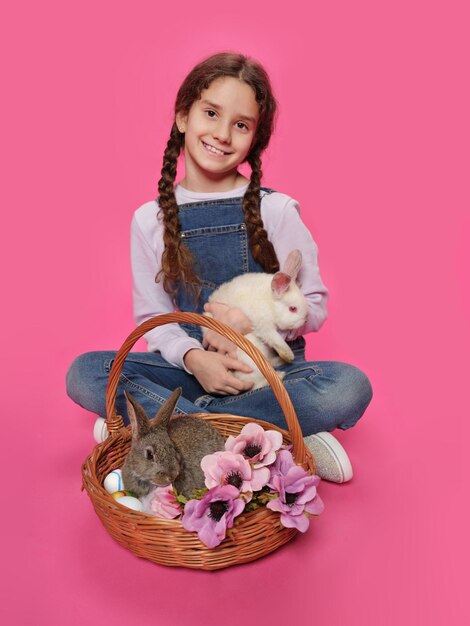 Vue pleine longueur d'une adorable petite fille assise sur le sol tenant dans les mains un petit lapin et un œuf de Pâques isolé sur fond rose Vue verticale