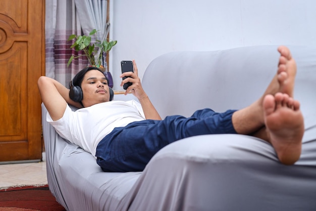 Vue pleine longueur d'un homme asiatique avec smartphone et casque posé sur le canapé à la maison