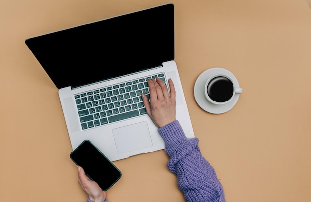 Vue POV sur une main féminine sur un ordinateur portable et tient un téléphone portable à côté d'une tasse de café sur fond marron