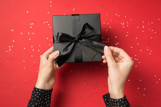 Vue à la première personne photo aérienne d'une boîte-cadeau noire avec un ruban enveloppé comme un arc dans les mains et des confettis autour isolés sur le fond rouge