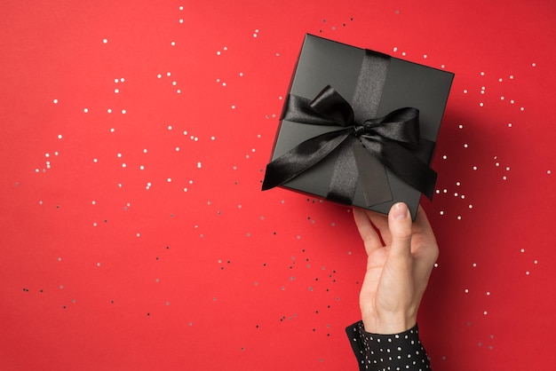 Vue à la première personne photo aérienne d'une boîte-cadeau noire avec un ruban enveloppé comme un arc dans les mains d'une femme et des confettis autour isolés sur le fond rouge