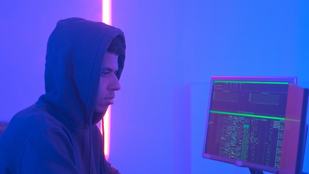 Vue de profil d'un hacker africain en sweat à capuche travaillant dans une pièce remplie d'écrans d'affichage et de couleurs chaudes ...