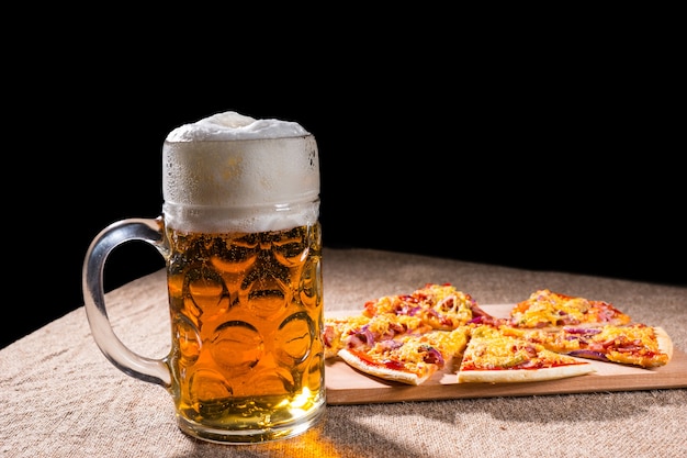 Vue de profil latéral d'une chope de bière avec une tête mousseuse à côté de tranches de pizza disposées sur une planche à découper en bois sur une surface de table recouverte de toile de jute