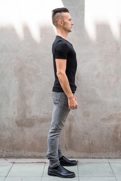 Vue de profil pleine longueur coup de beau jeune homme debout contre un mur gris à l'extérieur