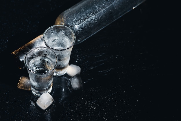 Vue rapprochée de la bouteille de vodka avec des verres debout sur la glace sur fond noir