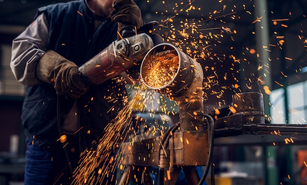 Vue rapprochée de côté de l'homme travailleur professionnel concentré en uniforme travaillant sur la sculpture de tuyau métallique avec une meuleuse électrique tandis que des étincelles volent dans l'atelier de tissu industriel.