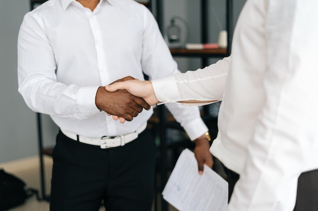 Vue rapprochée de divers partenaires commerciaux se serrant la main en accord debout dans la salle de bureau. Collègues afro-américains et caucasiens portant des vêtements de cérémonie se serrant la main et discutant d'affaires