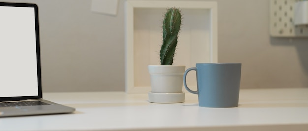 Vue rapprochée du bureau à domicile avec pot de cactus, tasse, cadre et maquette d'ordinateur portable sur un bureau blanc