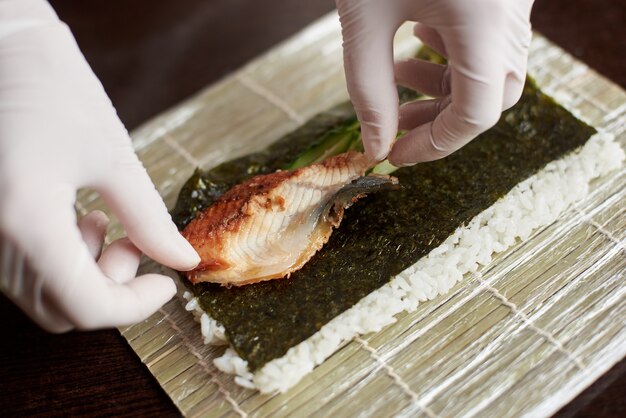 Vue rapprochée du processus de préparation des sushis roulants. Nori, riz blanc, concombre et anguille sur tapis de bambou. Les mains du chef dans les gants commencent à cuisiner des rouleaux de sushi