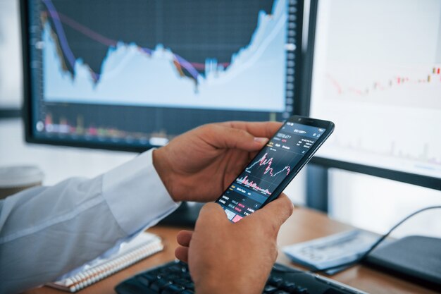 Vue rapprochée du téléphone tient par les mains de l'homme. L'homme d'affaires est au bureau avec plusieurs écrans. Conception de l'échange et de la monnaie.