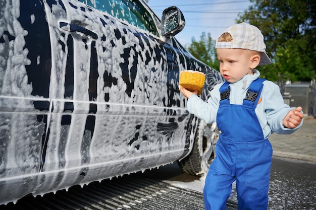 Vue rapprochée d'un enfant en bas âge avec une expression faciale sérieuse debout à côté d'une voiture noire recouverte de mousse et regardant l'automobile dans la confusion Petit garçon apprenant une nouvelle leçon