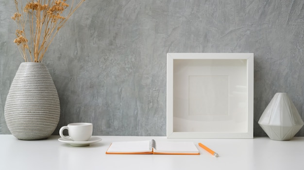 Vue rapprochée de l'espace de travail avec cahier ouvert, tasse à café, cadre et vases en céramique sur tableau blanc avec mur loft gris