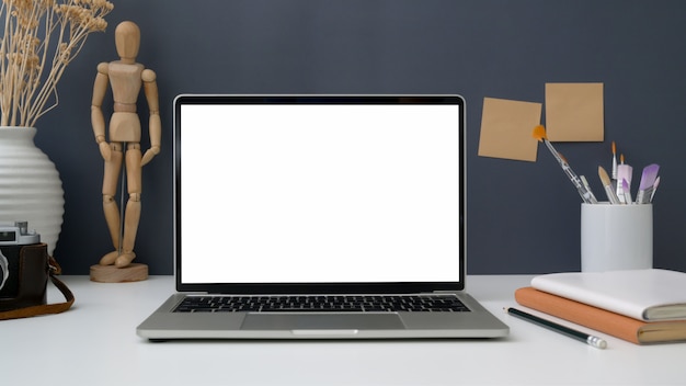 Vue rapprochée de l'espace de travail avec un ordinateur portable à écran blanc ouvert, des outils de peinture et des fournitures de bureau sur un tableau blanc avec mur gris