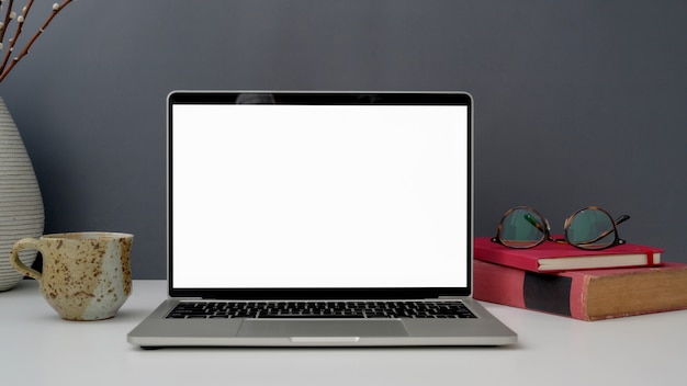 Vue rapprochée de l'espace de travail avec un ordinateur portable à écran blanc ouvert, une tasse, des verres et des livres sur un tableau blanc avec mur gris