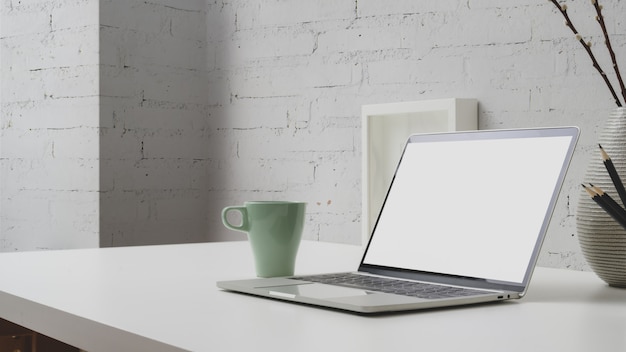 Vue rapprochée de l'espace de travail avec un ordinateur portable à écran blanc, une tasse de café et des décorations sur un bureau avec un mur de briques