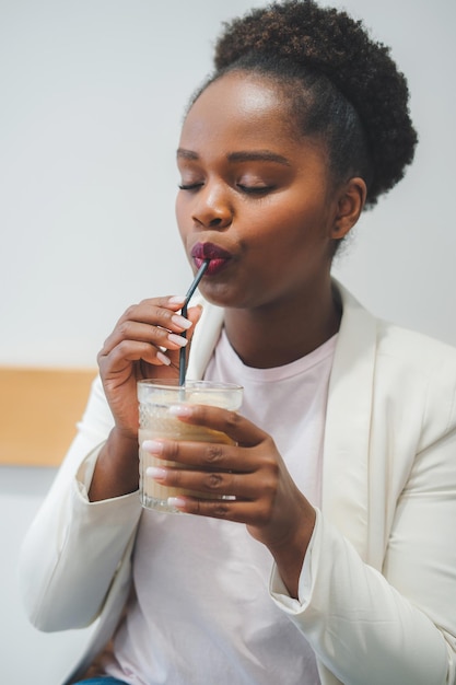 Vue rapprochée d'une femme africaine assise dans un café et buvant du café frappé en profitant de la pause-repas Concept de boissons et de personnes Beauté naturelle Portrait de beauté Mur blanc