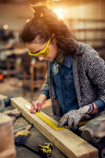 Vue Rapprochée De La Femme Charpentière Sérieuse Professionnelle Travailleuse Concentrée Tenant La Règle Et Le Crayon Tout En Faisant Des Marques Sur Le Bois à La Table Dans L'atelier De Tissu.