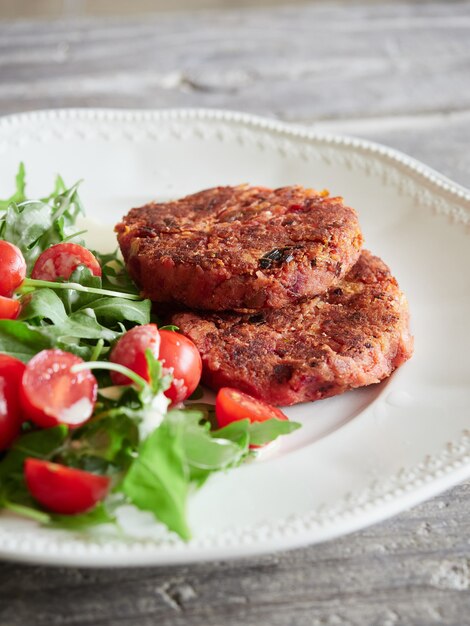 Vue rapprochée des hamburgers végétariens et de la salade de tomates cerises à la roquette servis sur une plaque blanche sur une table en bois. Alternative à la viande.