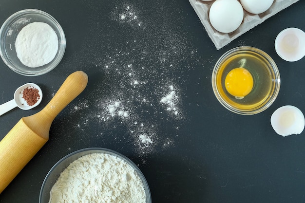 Vue rapprochée des ingrédients pour la cuisson d'un gâteau et d'un rouleau à pâtisserie en bois sur fond noir. Orientation horizontale, mise au point sélective. Copiez l'espace.